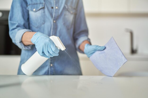 Primer plano de manos femeninas en guantes de goma con detergente en aerosol y paño durante la limpieza