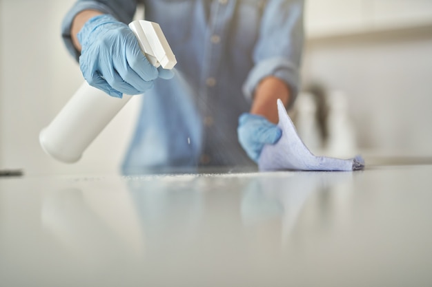 Primer plano de manos femeninas en guantes de goma con detergente en aerosol y paño mientras limpia blanco