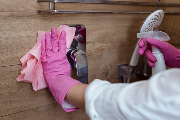 Primer plano de manos femeninas con guantes de goma lavando la cabina de ducha y el grifo en el baño