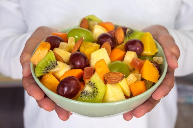 Primer plano de manos femeninas sosteniendo un tazón lleno de fruta fresca y colorida Manzana kiwi naranja papaya uva mango melón almendras Desayuno o descanso concepto de alimentación saludable