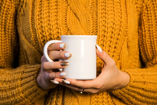 Primer plano de manos femeninas con una taza de bebida hermosa mujer en gritar suéter sosteniendo una taza de té