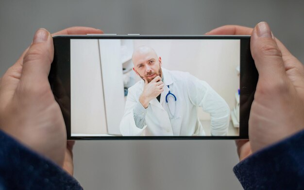 Primer plano de manos femeninas con un teléfono Un médico calvo consulta de forma remota a un paciente con una cámara web La mujer dice con un médico amigable para videollamadas