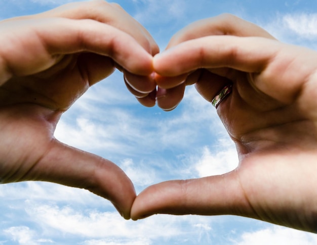 Primer plano de manos en forma de corazón contra el cielo azul con nubes blancas.