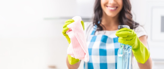 Primer plano de manos con guantes de goma amarillos sosteniendo un detergente y un trapo de limpieza por una mujer sonriente borrosa en la espalda con un delantal a cuadros azul y blanco