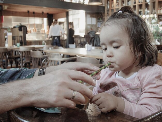 Foto primer plano de manos de un hombre ayudando a una niña a beber jugo de un vaso en la mesa de un restaurante