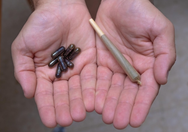 Primer plano de las manos de un hombre sosteniendo pastillas en una y un porro de cannabis en la otra