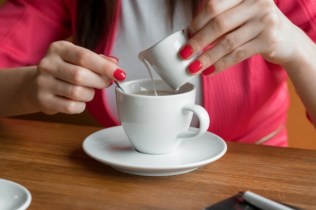 Primer plano, las manos de una joven, vierte crema o leche en el café en un café en la mesa de madera.