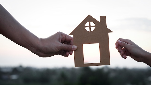 Primer plano de las manos de madre e hija sosteniendo la casa de papel juntos contra la puesta de sol con enfoque suave