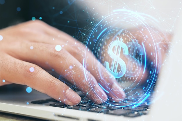 Un primer plano de manos masculinas usando una computadora portátil con un holograma creativo de dólares en un fondo borroso del escritorio de la oficina.