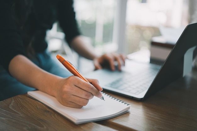 Primer plano de las manos de una mujer de negocios que trabaja en una computadora portátil y usa un bolígrafo para hacer notas en su cuaderno en una mesa de madera con una taza de café Concepto de negocio