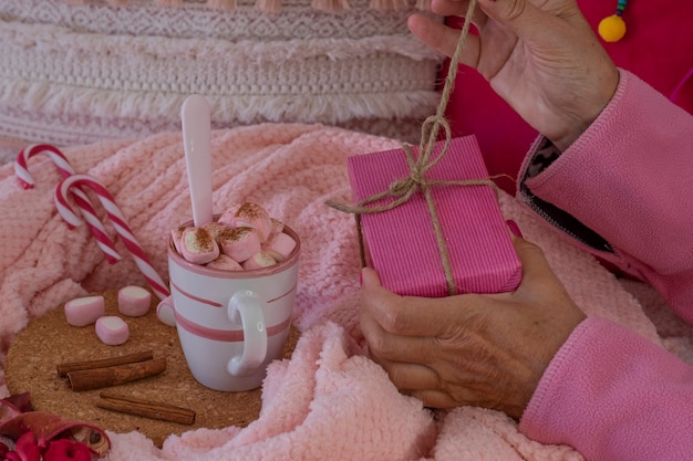 Primer plano de las manos de una mujer en pijama rosa abriendo un paquete de regalo, cerca de ella una taza con chocolate caliente con malvaviscos y canela en polvo. Vacaciones de Navidad y concepto de gente feliz