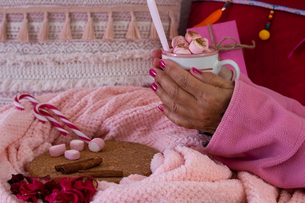 Primer plano de las manos de una mujer en pijama rosa sosteniendo un chocolate caliente con malvaviscos en una taza de cerámica. Concepto de vacaciones y personas