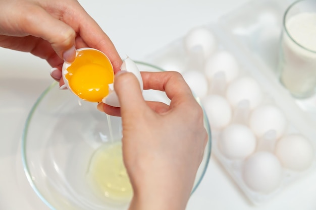 Primer plano de las manos de una mujer que rompe un huevo con los dedos y vierte la yema en un plato contra el fondo de un paquete con una docena de huevos y un vaso con harina