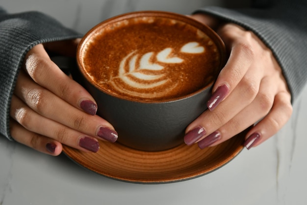 Primer plano de las manos de una mujer sosteniendo una taza de café americano con espuma espesa, bebida de capuchino caliente,