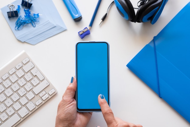 Primer plano de manos de mujer sosteniendo teléfono inteligente con pantalla azul. Color azul en accesorios.