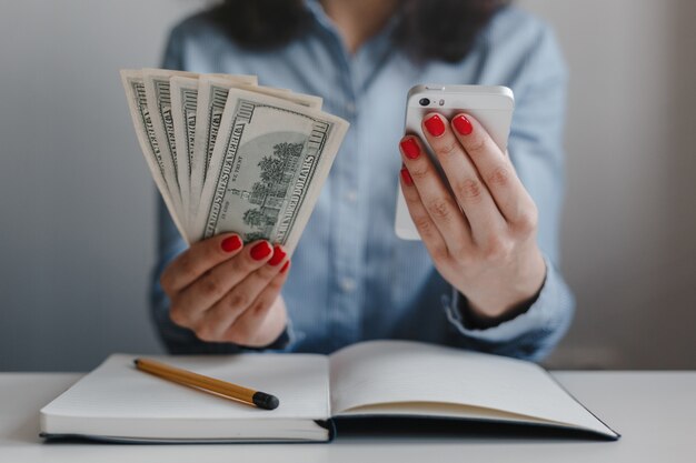 Primer plano de las manos de una mujer con uñas rojas sosteniendo billetes de cien dólares y un móvil