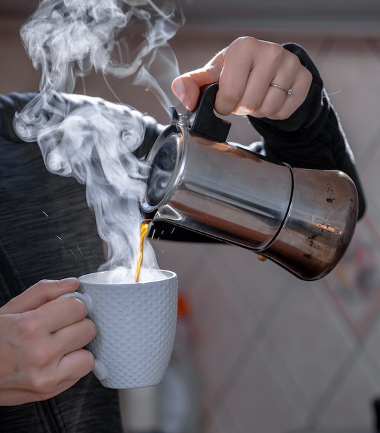 Primer plano de manos de mujeres adultas que vierten café caliente en una taza blanca de metal cafetera, el humo del café está humeando