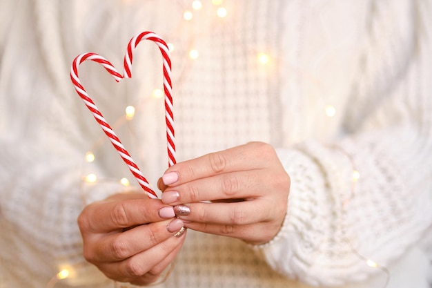 Primer plano de las manos de las mujeres sosteniendo bastones de caramelo en forma de corazón. Dulces en manos de una mujer con un suéter de punto blanco. Pequeña profundidad de campo, enfoque selectivo