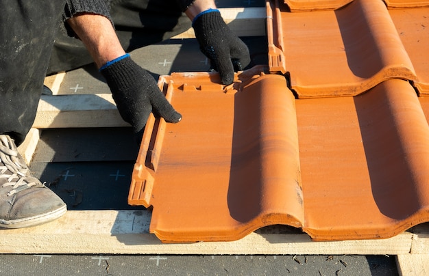 Primer plano de las manos del trabajador instalando tejas de cerámica amarillas montadas sobre tablas de madera que cubren el techo del edificio residencial en construcción.