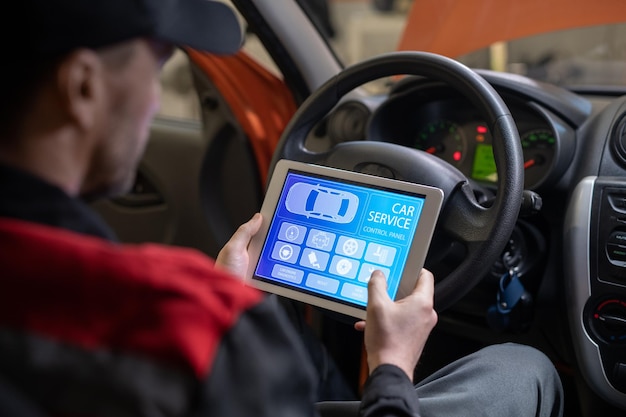 Primer plano del mecánico de automóviles que usa una tableta digital con la aplicación de servicio y mantenimiento en la pantalla mientras inspecciona el vehículo en el taller de reparación de automóviles