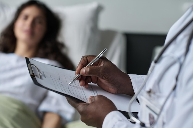 Primer plano de un médico afroamericano haciendo notas en la tarjeta médica mientras habla con el paciente en la sala