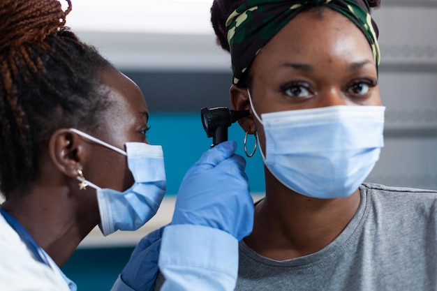 Primer plano del médico otólogo afroamericano mirando el oído del paciente enfermo con otoscopio médico durante el examen clínico en la oficina del hospital. Mujeres con mascarilla protectora contra el coronavirus.