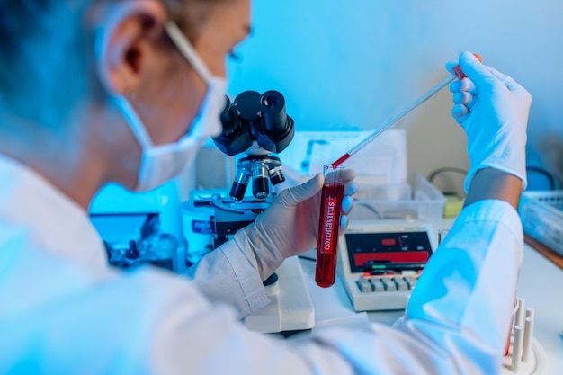 Foto primer plano: microscopio óptico de laboratorio. tubo de ensayo de diagnóstico en mano de mujer con guante blanco. análisis de muestra clínica de sangre. investigación científica del coronavirus.