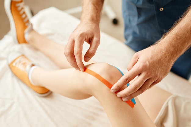Primer plano mínimo de fisioterapeuta poniendo cinta adhesiva en la rodilla de una mujer joven para aliviar el dolor muscular y articular
