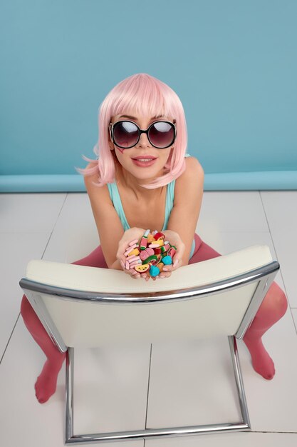 Primer plano de una modelo femenina coqueta sobre fondo azul con una peluca rosa sosteniendo diferentes tipos de dulces Mujer bastante glamorosa cabello rosa encanto dulces estilo de vida disfrute azúcar