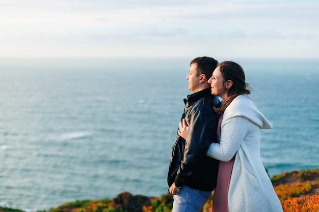 Un primer plano de una mujer abraza a su esposo desde la parte de atrás y admiran la vista al mar