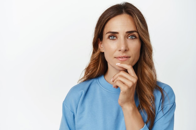 Primer plano de una mujer adulta reflexiva con una mirada sonriente y segura, tocar la barbilla, pensar, mirar a la cámara con cara de reflexión, tomar decisiones, elegir, pararse sobre fondo blanco.