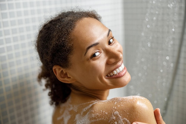 Primer plano de una mujer africana sonriente duchándose con gel o espuma de champú en el baño