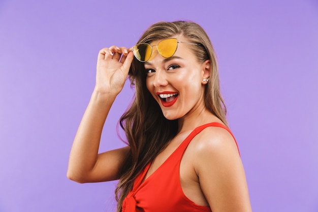 Primer plano de una mujer alegre y elegante con vestido rojo y gafas de sol sonriendo a la cámara