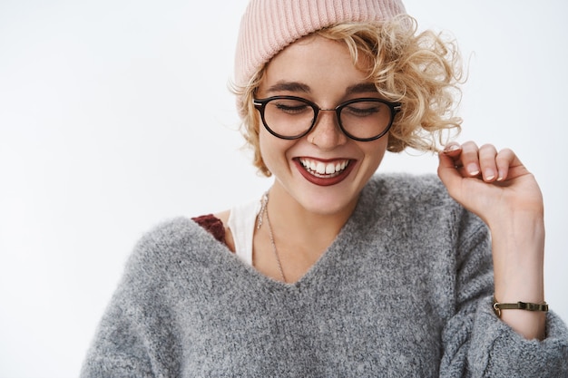 Primer plano de mujer alegre linda y tierna divertida con gorro de gafas y suéter de invierno cálido jugando con rizo de pelo mirando hacia abajo riendo tímido y despreocupado de felicidad y alegría, divirtiéndose