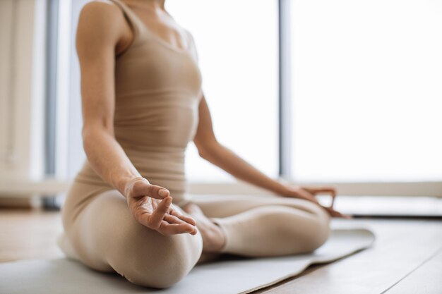 Un primer plano de una mujer delgada con una camiseta beige y pantalones de yoga logrando la posición del loto