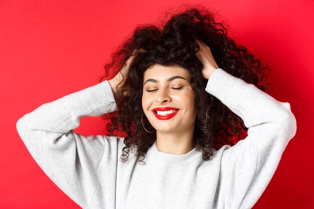 Primer plano de mujer despreocupada tocando el pelo rizado suave y sonriendo complacido, de pie sobre fondo rojo. Concepto de belleza y cuidado del cabello.