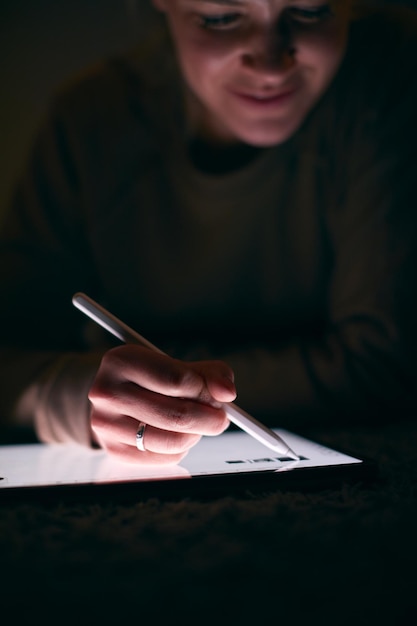 Primer plano de una mujer dibujando en una tableta digital usando un lápiz óptico recostado sobre una alfombra por la noche