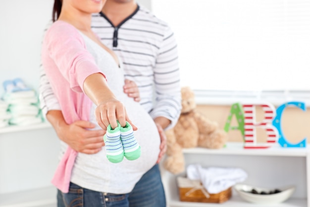 Primer plano de una mujer embarazada brillante con zapatos de bebé mientras el marido toca su vientre en la habitación de su futuro bebé