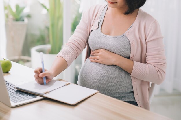 Primer plano de una mujer embarazada escribiendo notas y usando una computadora portátil mientras trabaja en la licencia de maternidad en la mesa de su comedor en casa
