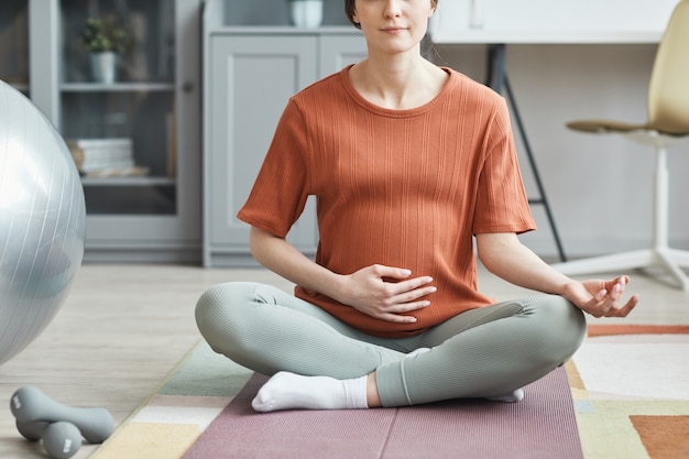 Primer plano de una mujer embarazada sentada en una colchoneta y meditando en la habitación de su casa