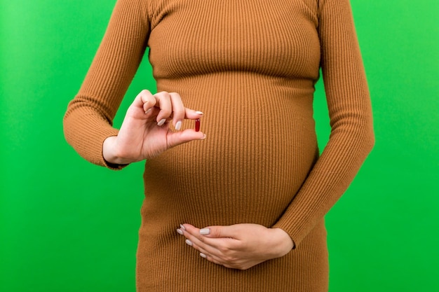 Primer plano de una mujer embarazada sosteniendo una píldora en la mano contra su vientre en un fondo colorido con espacio de copia Tomando vitaminas durante el embarazo concepto