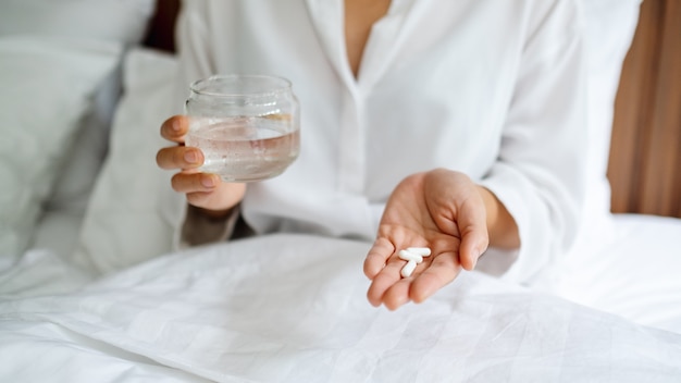 Foto primer plano de una mujer enferma con pastillas blancas y un vaso de agua mientras está sentado en una cama