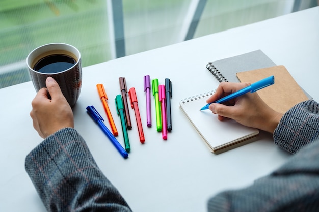 Primer plano de una mujer escribiendo en un cuaderno en blanco con bolígrafos de colores mientras bebe café