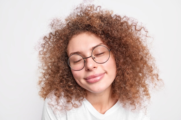 Primer plano de una mujer europea complacida con el pelo rizado y tupido que cierra los ojos por el placer y recuerda agradables recuerdos lleva gafas redondas transparentes aisladas sobre una pared blanca. Disfrutando la vida al máximo