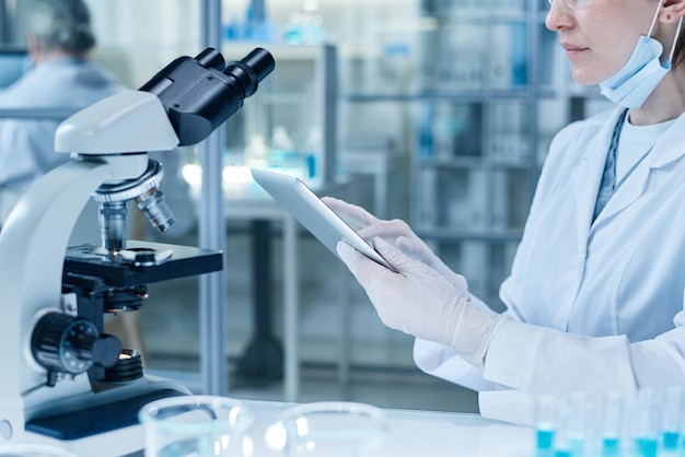 Primer plano de una mujer con guantes protectores y bata de laboratorio usando una tableta digital para examinar las muestras en la pantalla después de investigarlas en el microscopio