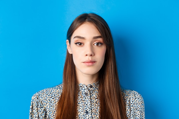 Primer plano de mujer hermosa sincera con maquillaje y cabello largo natural, mirando a cámara, de pie en azul.