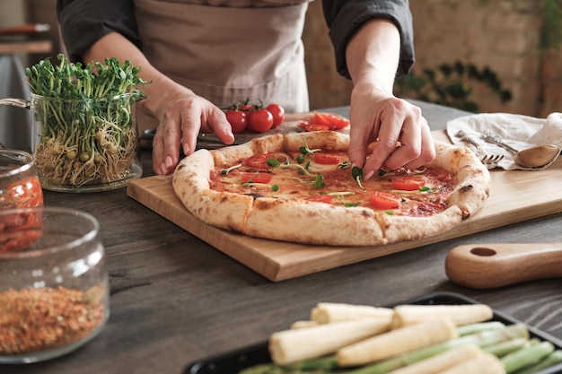 Primer plano de una mujer irreconocible en delantal de pie en la mesa con frascos de especias y decorando pizza caliente con hierbas