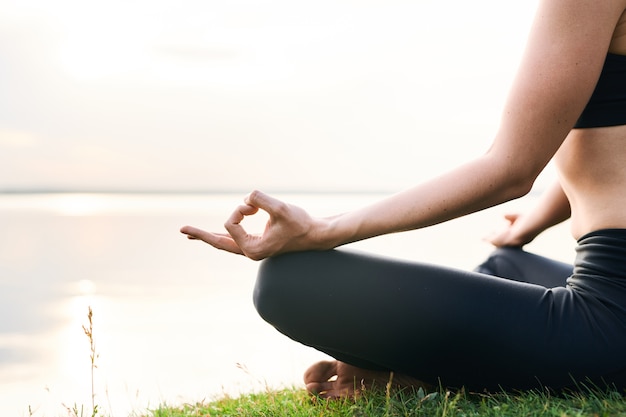 Primer plano de mujer irreconocible en leggings sentada en la orilla del lago y practicando yoga en unidad con la naturaleza