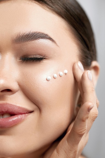 Primer plano mujer joven feliz aplicando crema en la cara Concepto de cuidado de la piel y cosméticos Cosméticos
