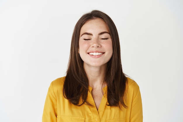 Primer plano de mujer joven feliz y encantada sonriendo con los ojos cerrados, pidiendo un deseo o soñando, de pie en la pared blanca